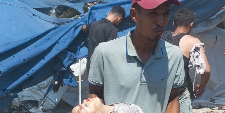 A man holding a dead child in Gaza amid rubble and blue tents.