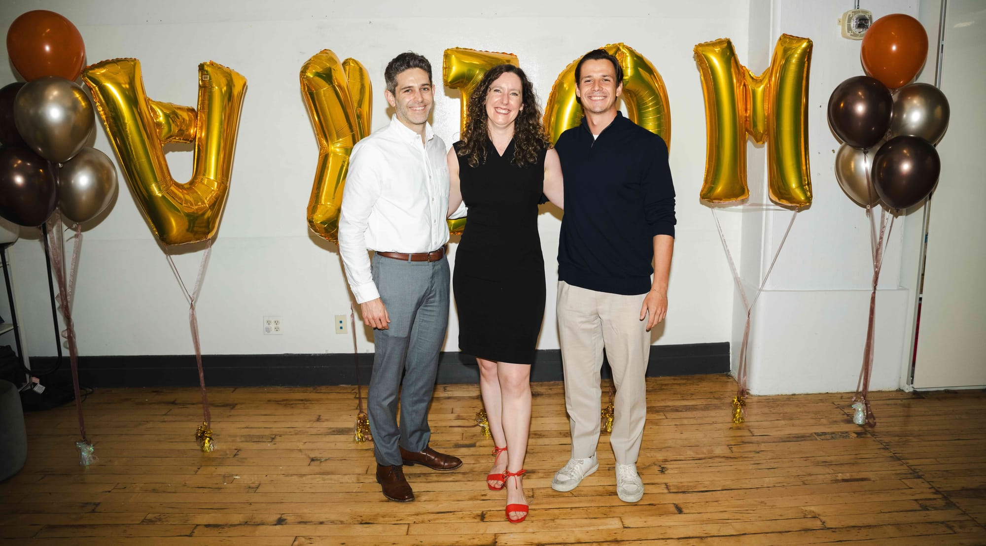Brooklyn loft with wooden floors, two clusters of bronze and orange balloons and gold letter balloons spelling AXIOM. The founding team of the Axiom Foundation stands in front, Max in a white shirt and gray pays, Ariel in a black dress and red sandals, Pavel in an navy sweater and tan pants. 