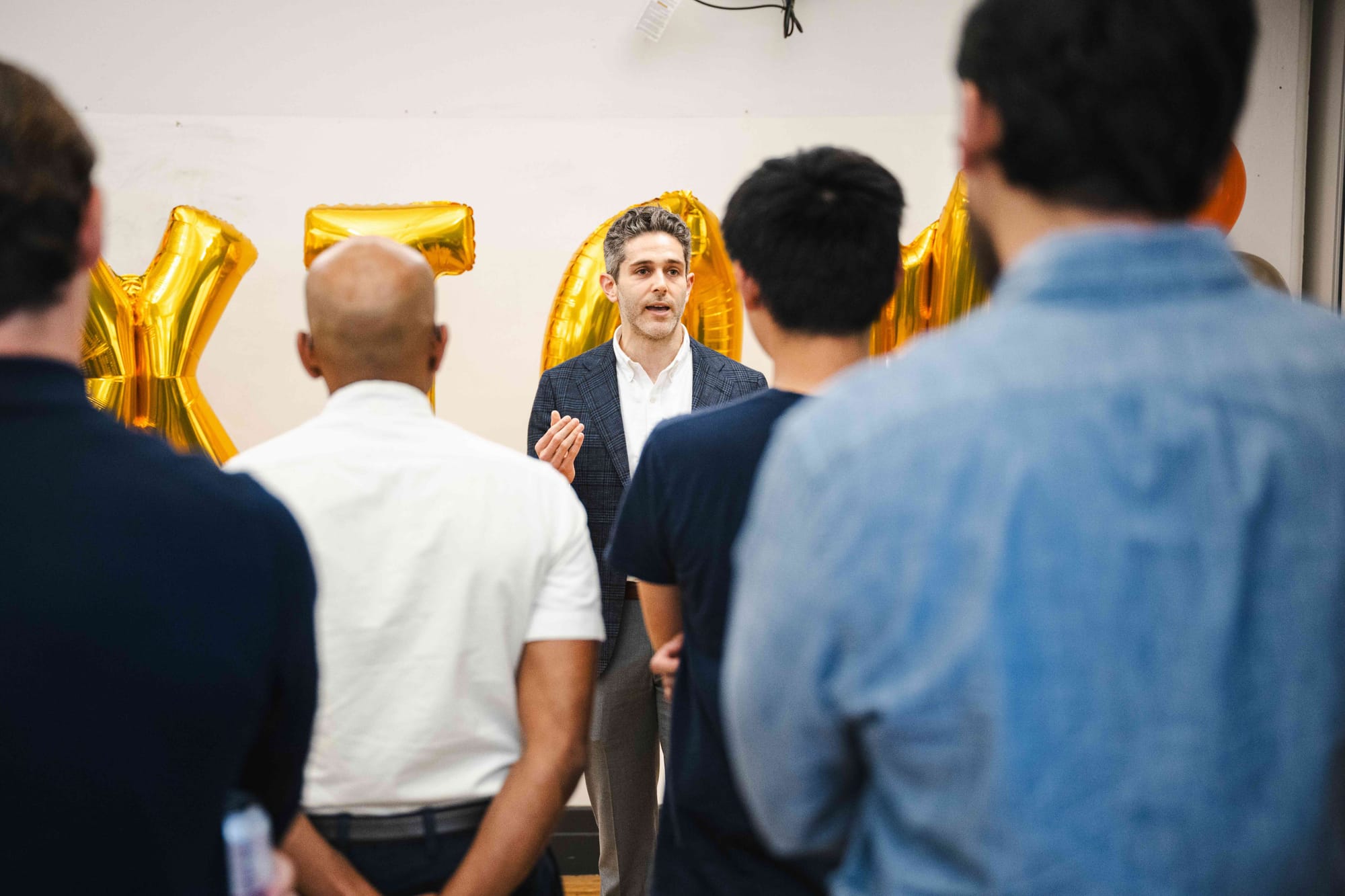 Max Ghenis speaking to guests in front of the gold AXIOM balloons, listeners in the foreground.
