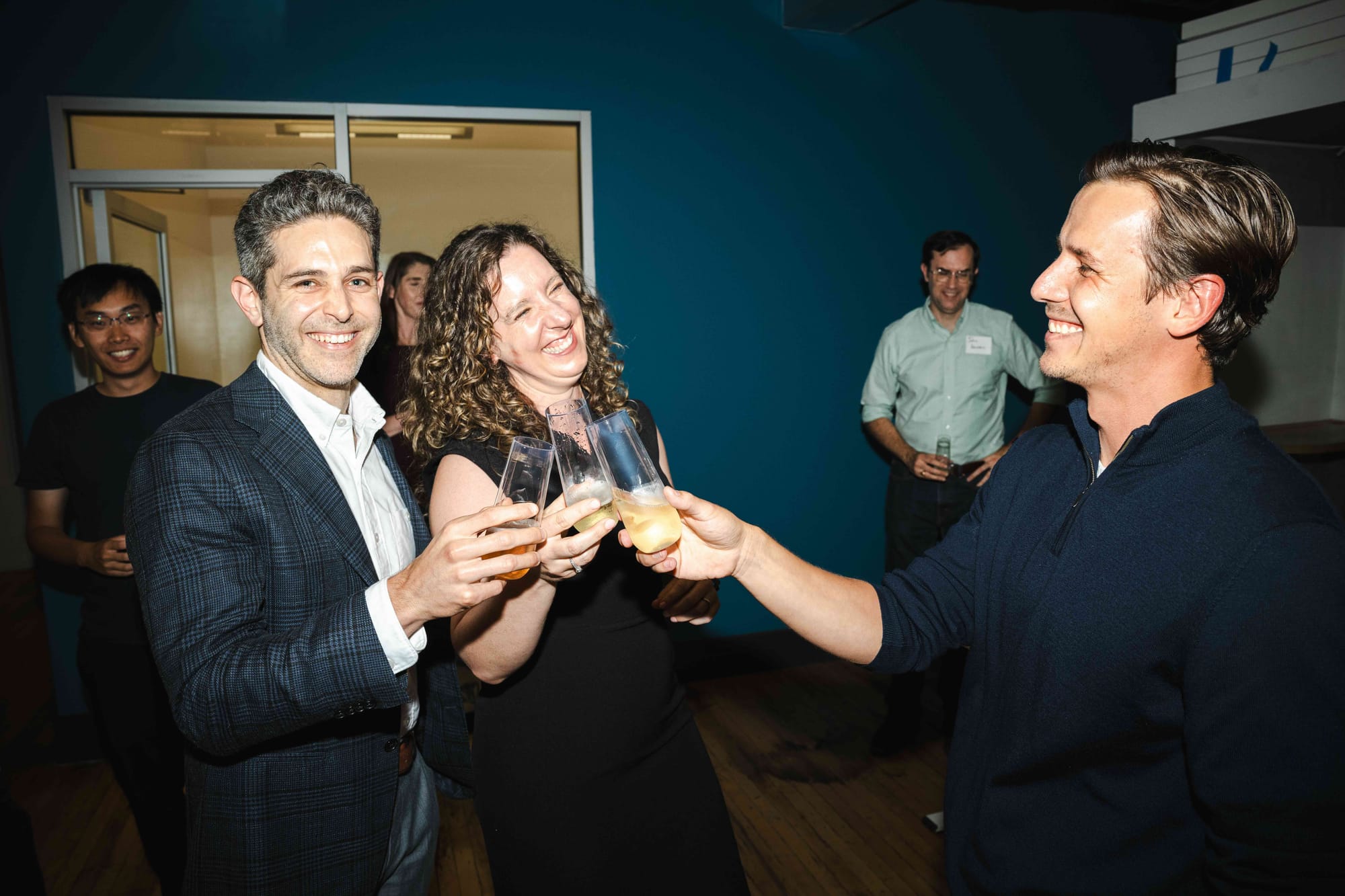 Max Ghenis, Ariel Kennan, and Pavel Makarchuk clinking glasses in a toast, laughing, against a deep blue wall.