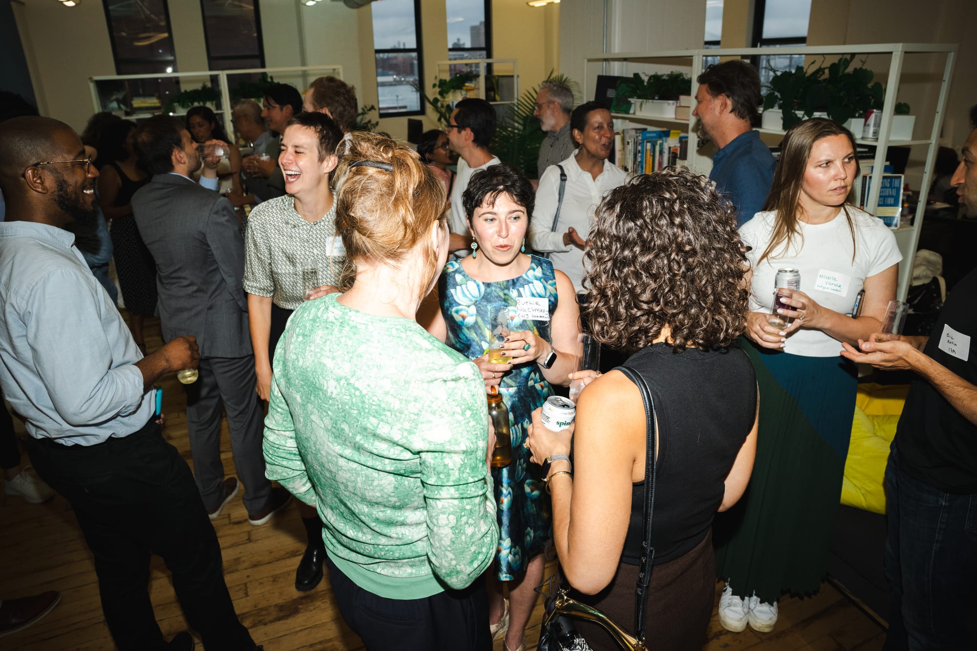 Guests in conversation clusters at the reception, name tags on, with bookshelves and evening city windows behind.