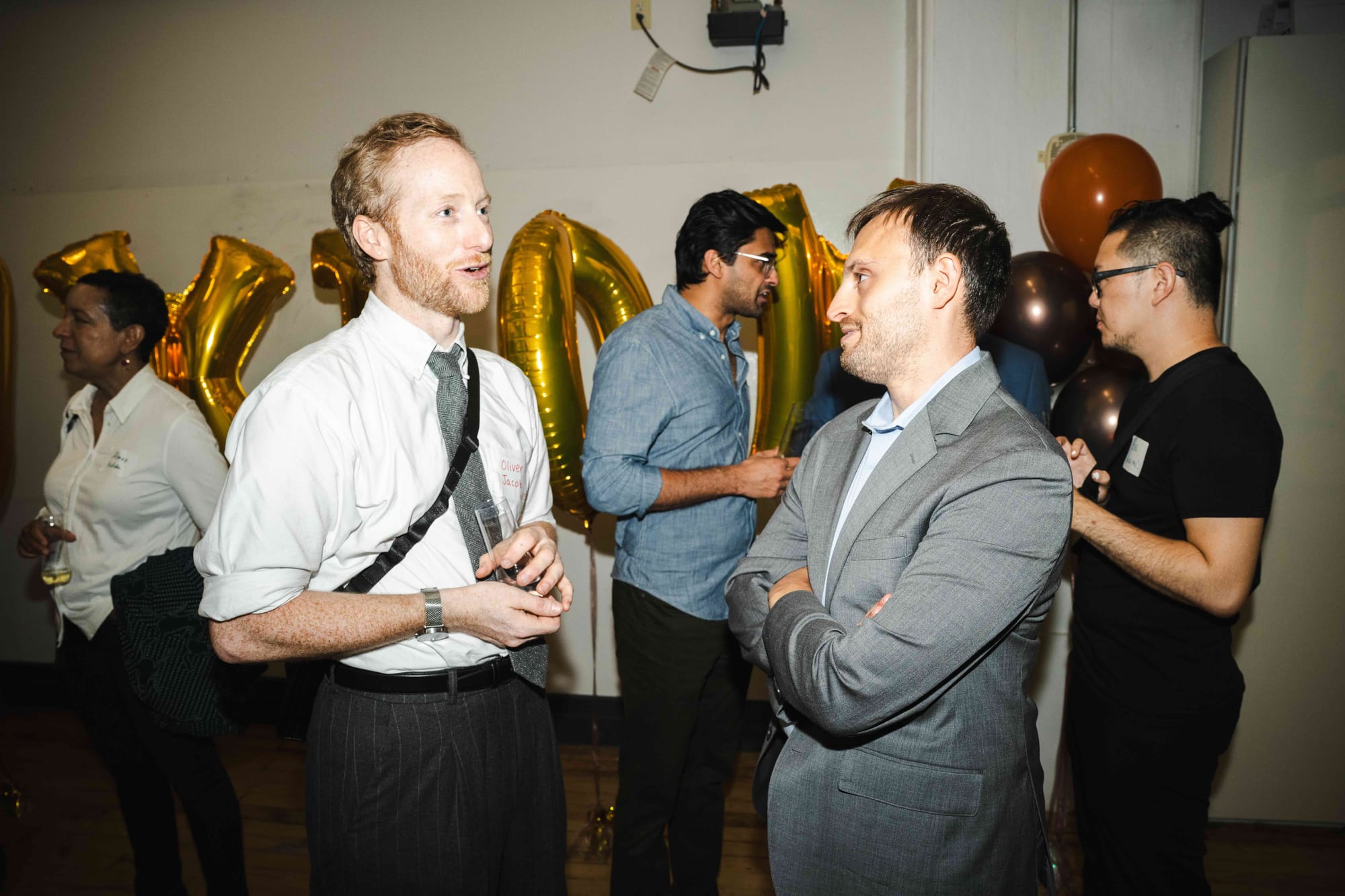 Two guests deep in conversation at the reception, with more small groups and balloons behind them.