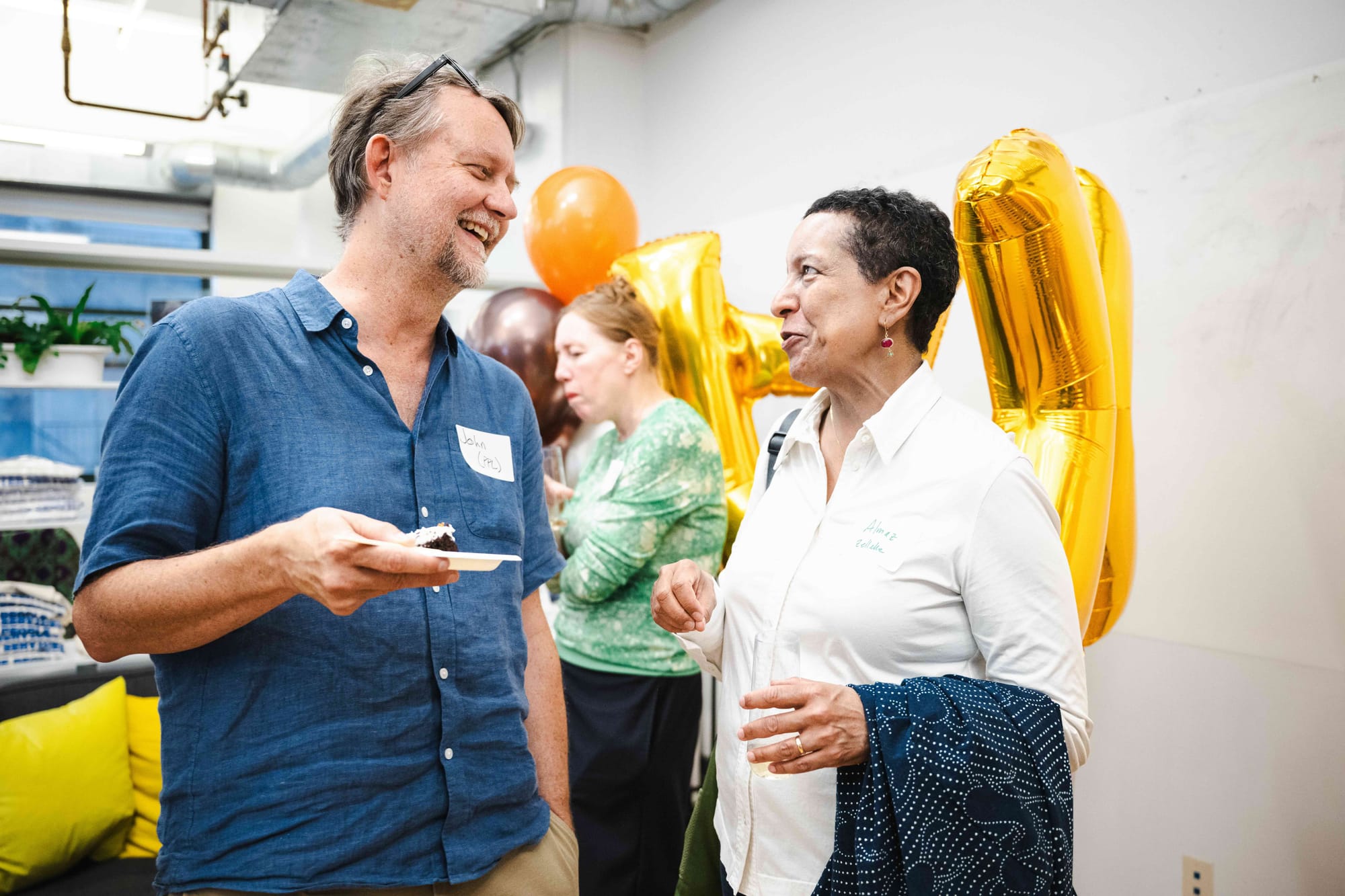 Two guests laughing mid-conversation, one holding a slice of cake, in front of gold letter balloons.