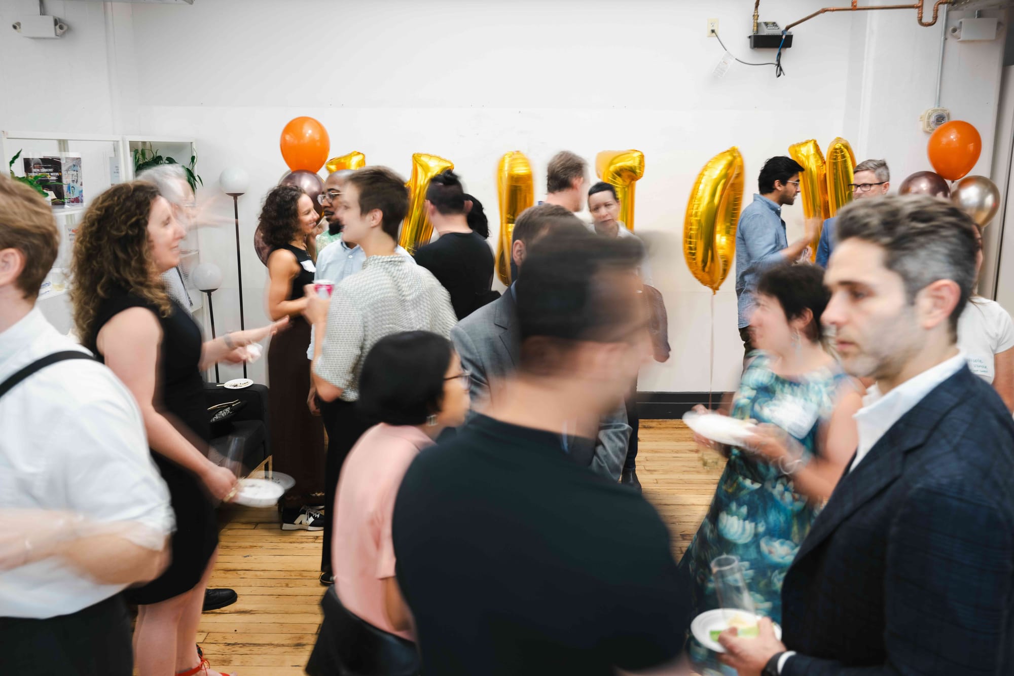 A crowded loft reception in motion, guests holding plates and drinks in front of gold foil balloons spelling AXIOM.