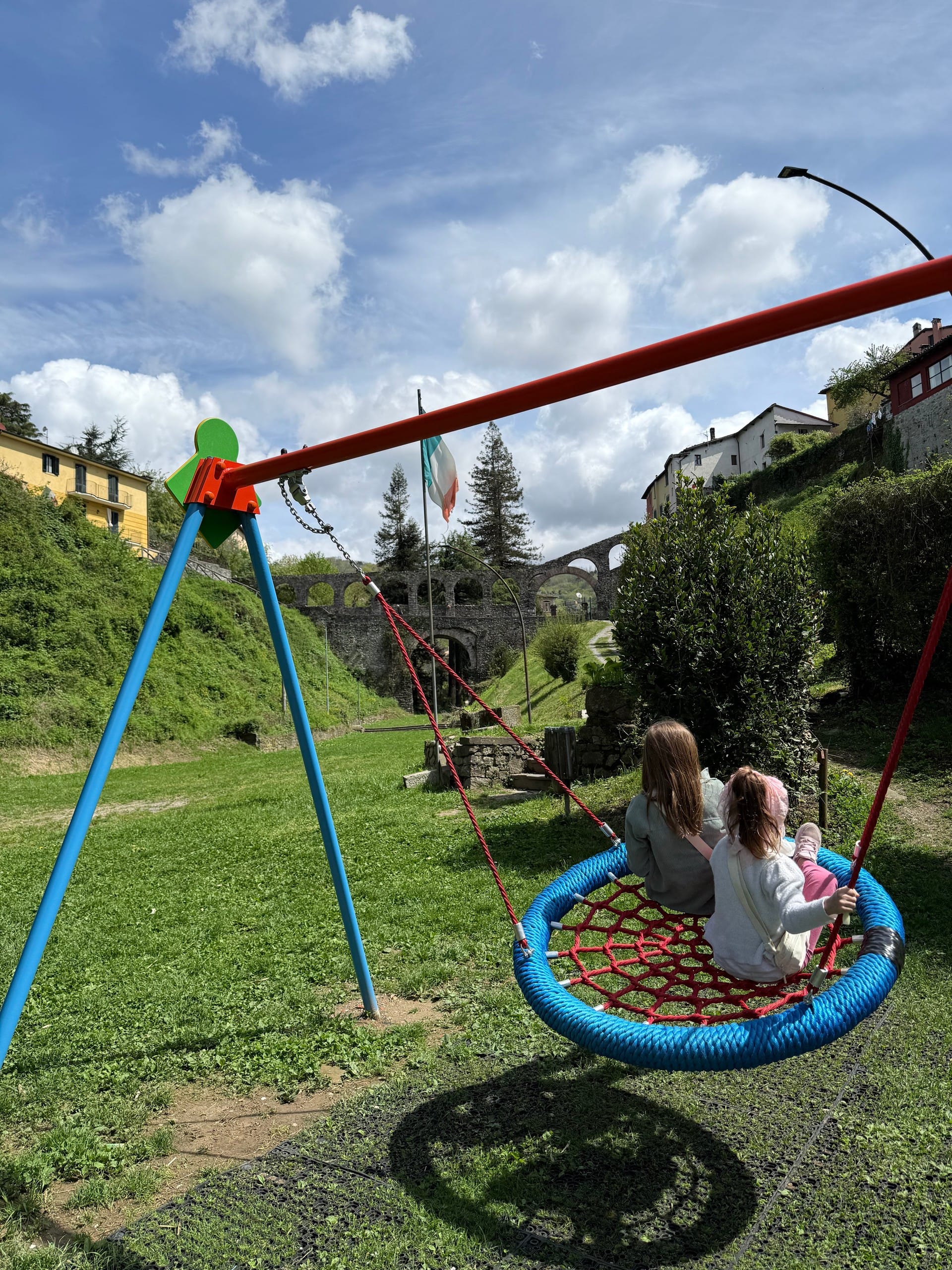 Lucy and Izzy on a swing set in a Barga, Italy valley, with a stone bridge in the background