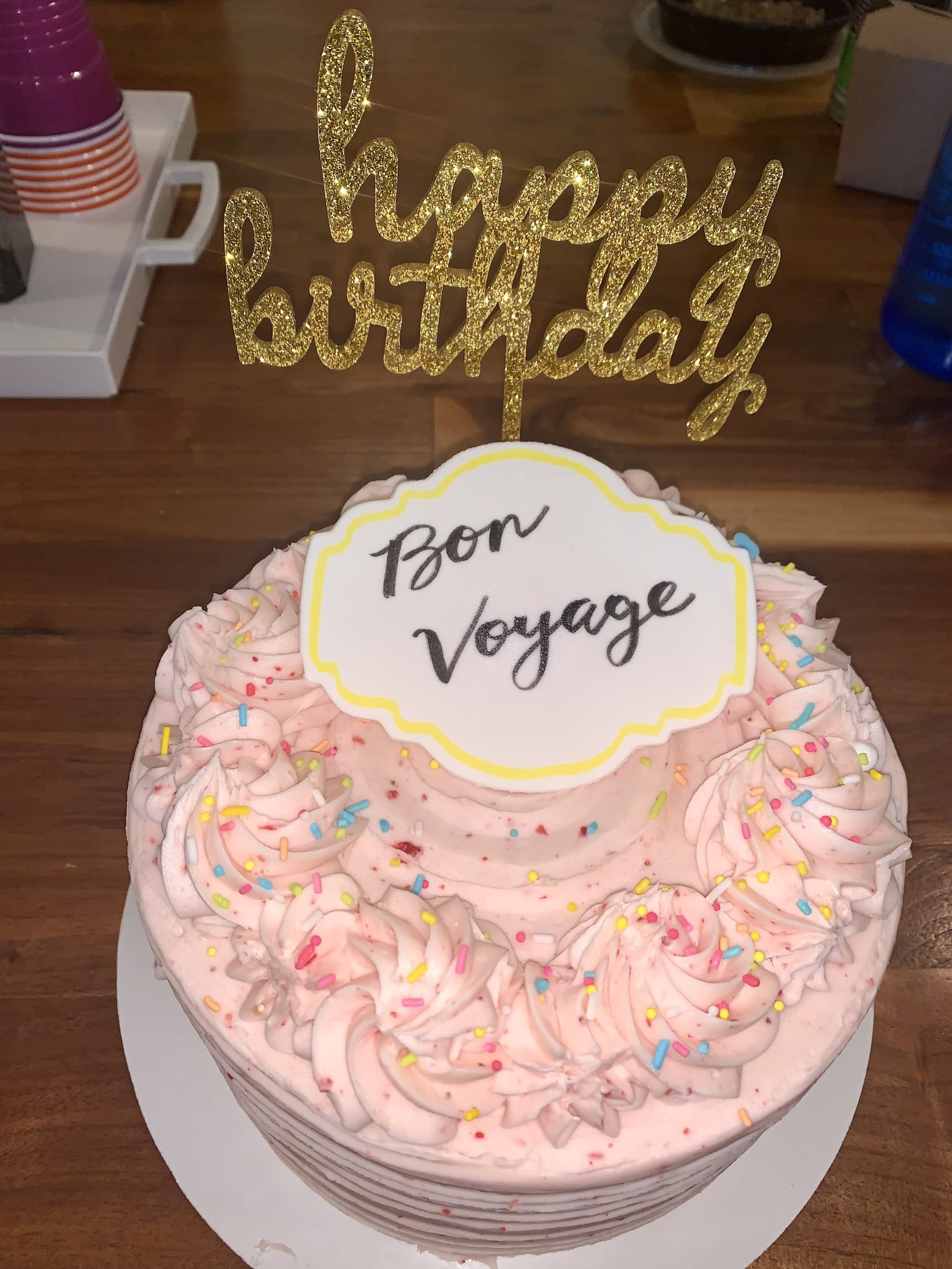 A pink frosted cake with a gold "Happy Birthday" topper and a "Bon Voyage" plaque, set out for a going-away celebration.