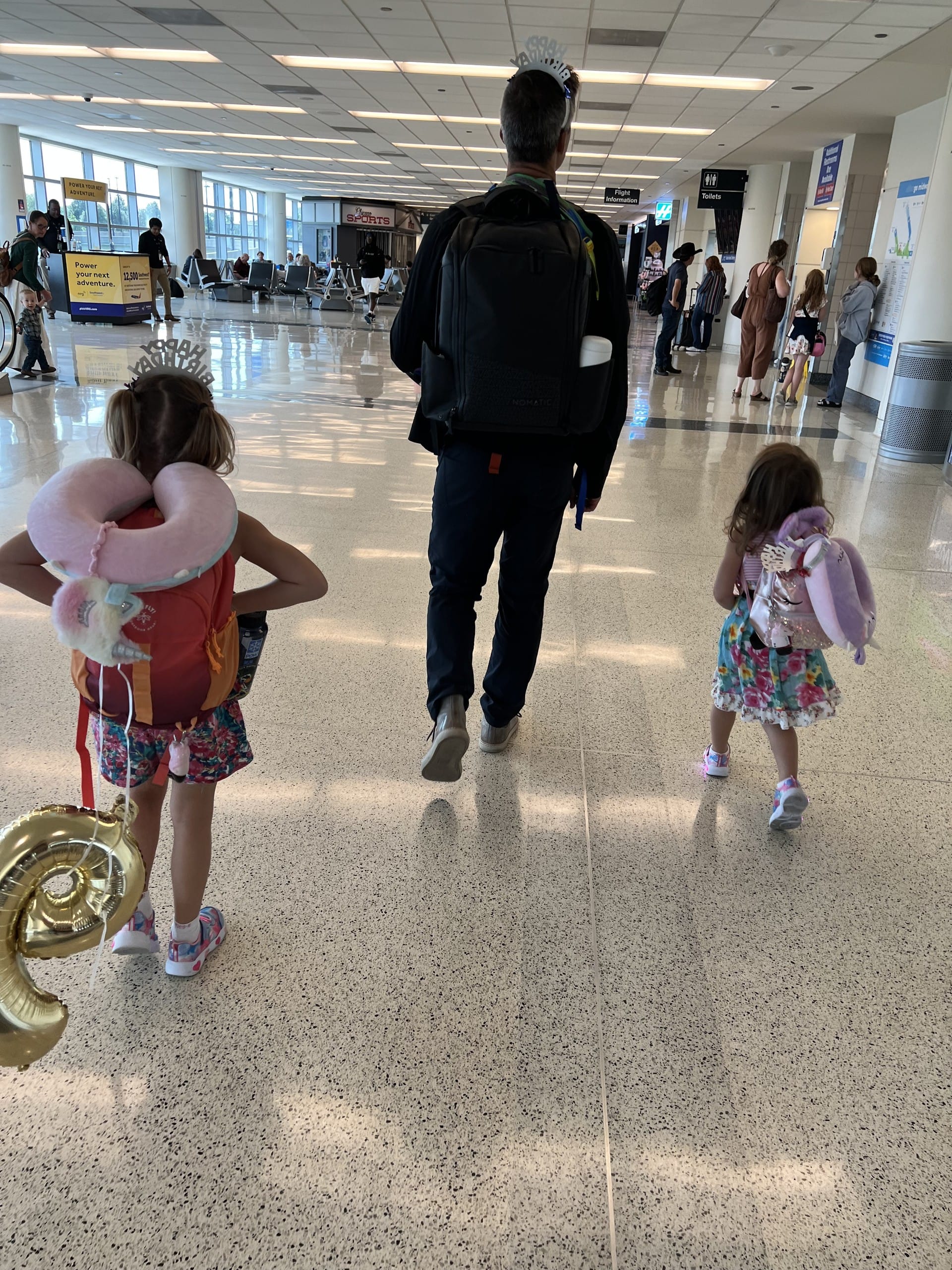 Dad and two girls walking through the airport concourse, backs to camera, a deflating number-6 balloon in tow