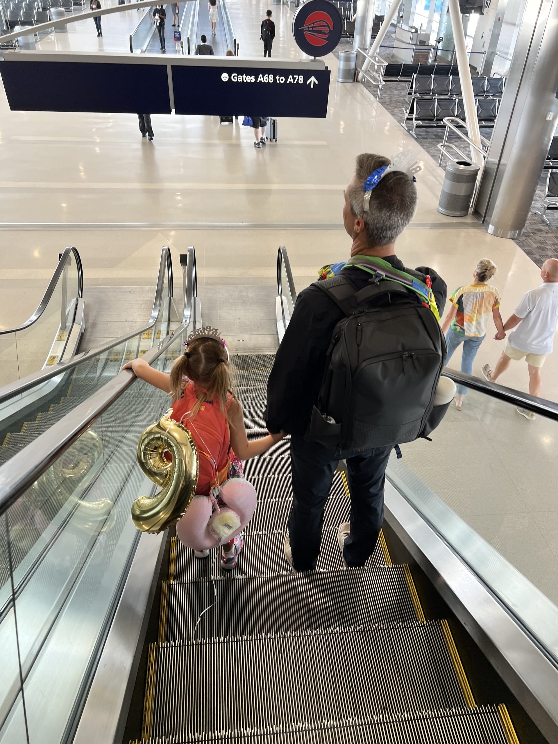 Dad and daughter riding an airport escalator toward the gates, balloon in hand