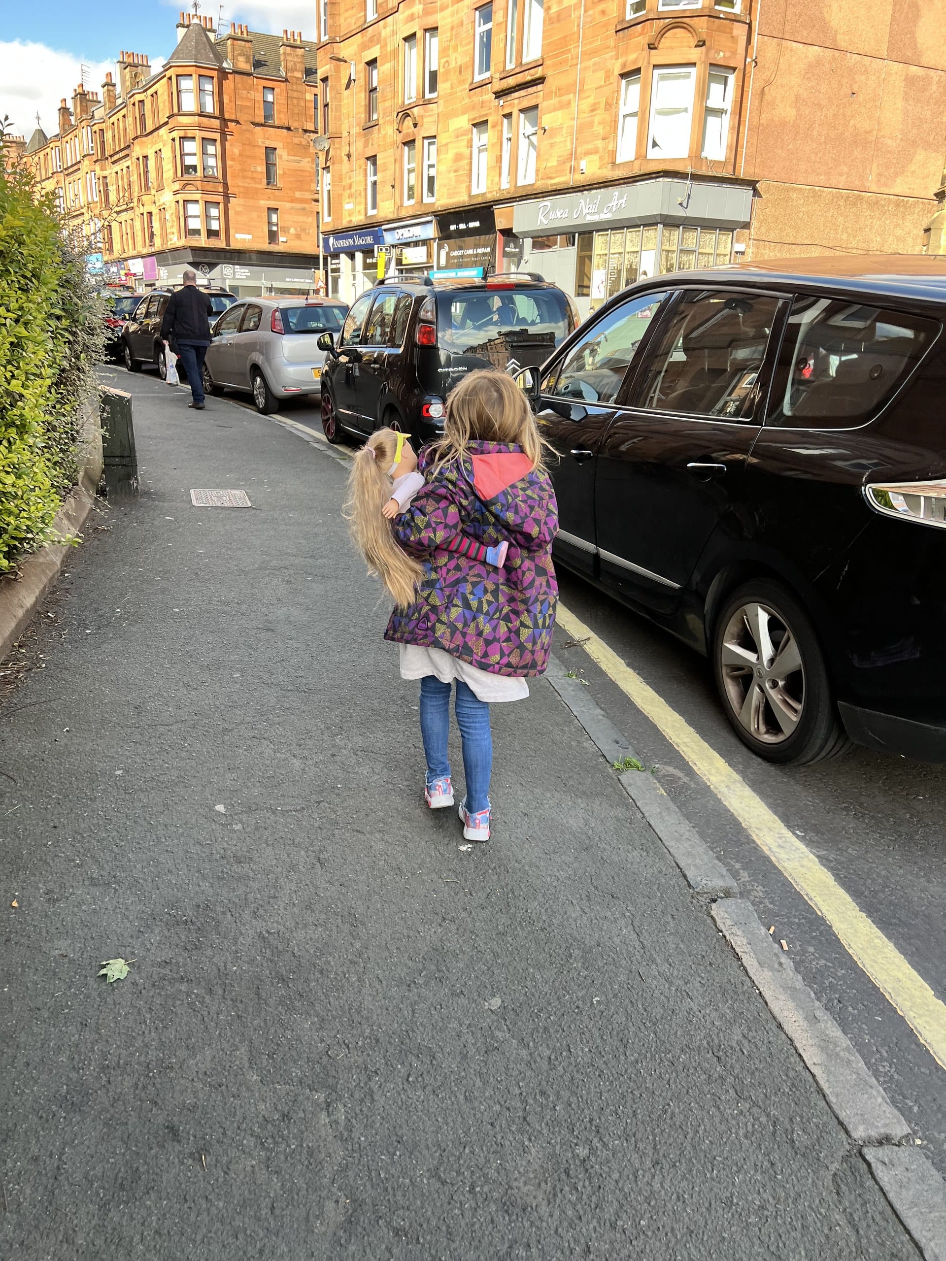 A girl carries her doll along a residential street lined with shops and tenements in Mount Florida, Glasgow