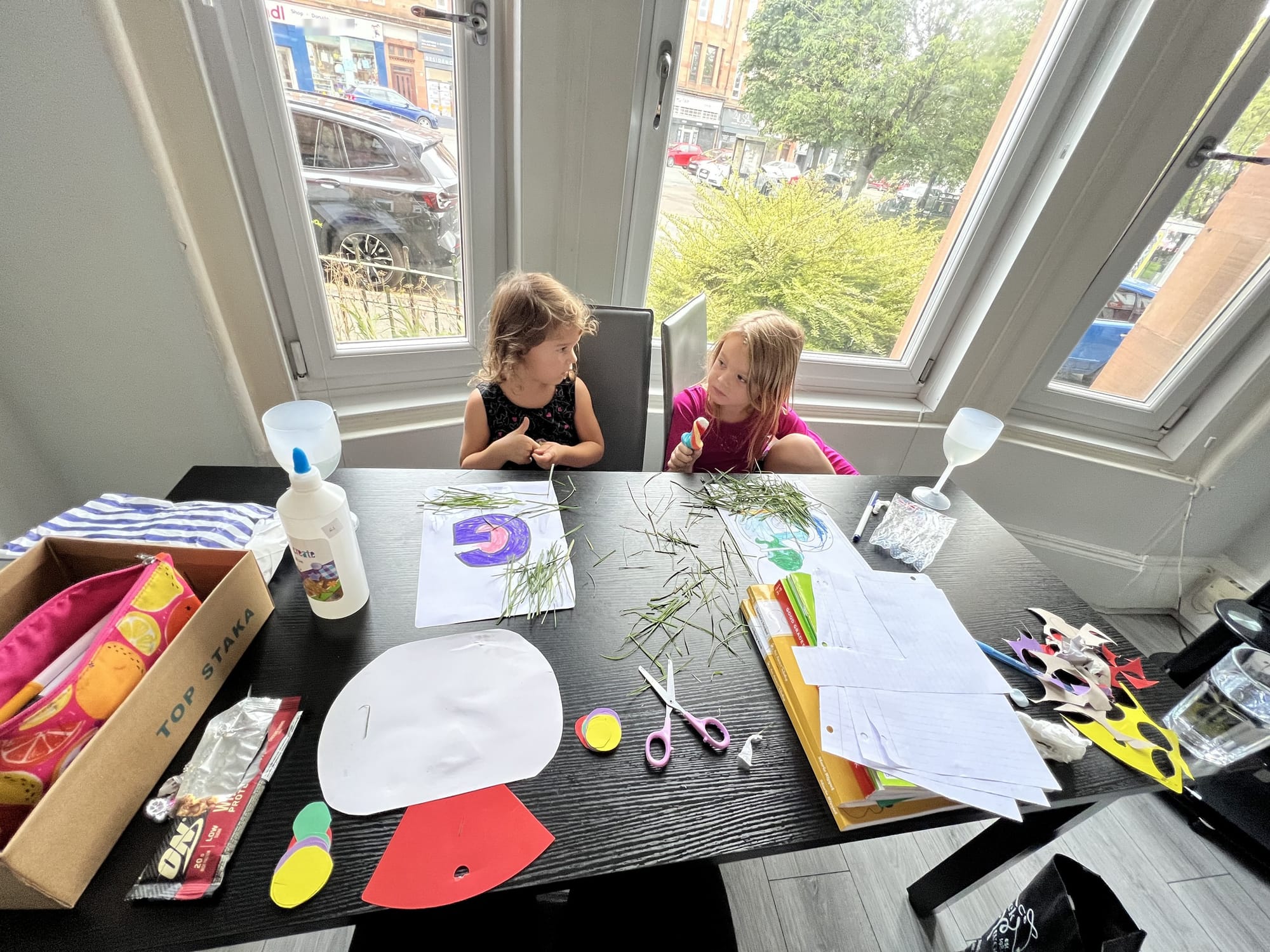 Two girls doing schoolwork at a bay-window table, Glasgow tenement buildings visible through the window.