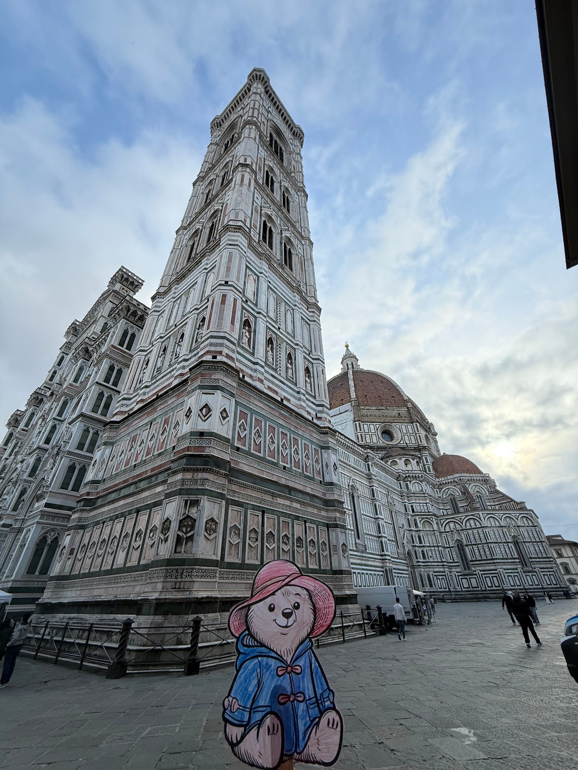 A child's own hand-drawn, hand-colored paper bear in a rose-pink hat, no lettering, propped up at the foot of Giotto's Campanile, Florence, with Brunelleschi's dome behind.