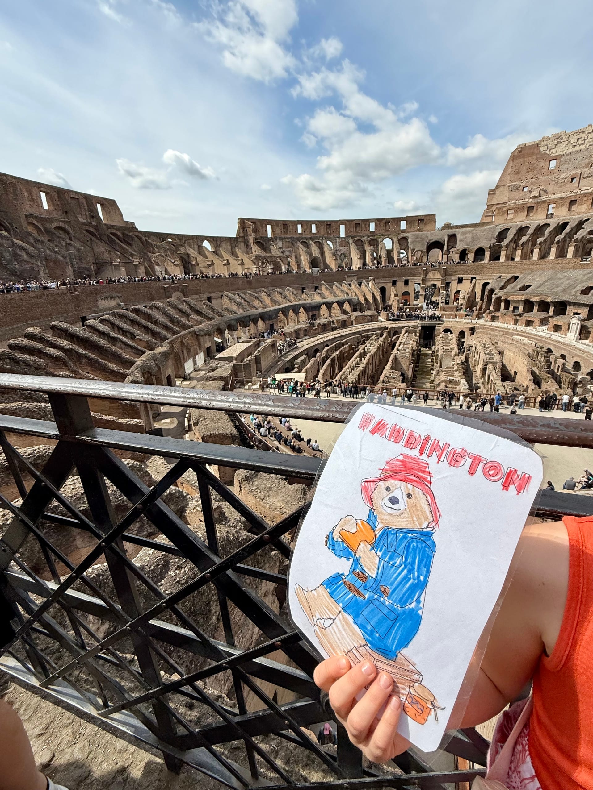 A child's hand holding a laminated, hand-colored paper bear in a red hat, its name lettered across the top in the child's own handwriting, held up over a railing inside the Colosseum, the arena floor and passages below.