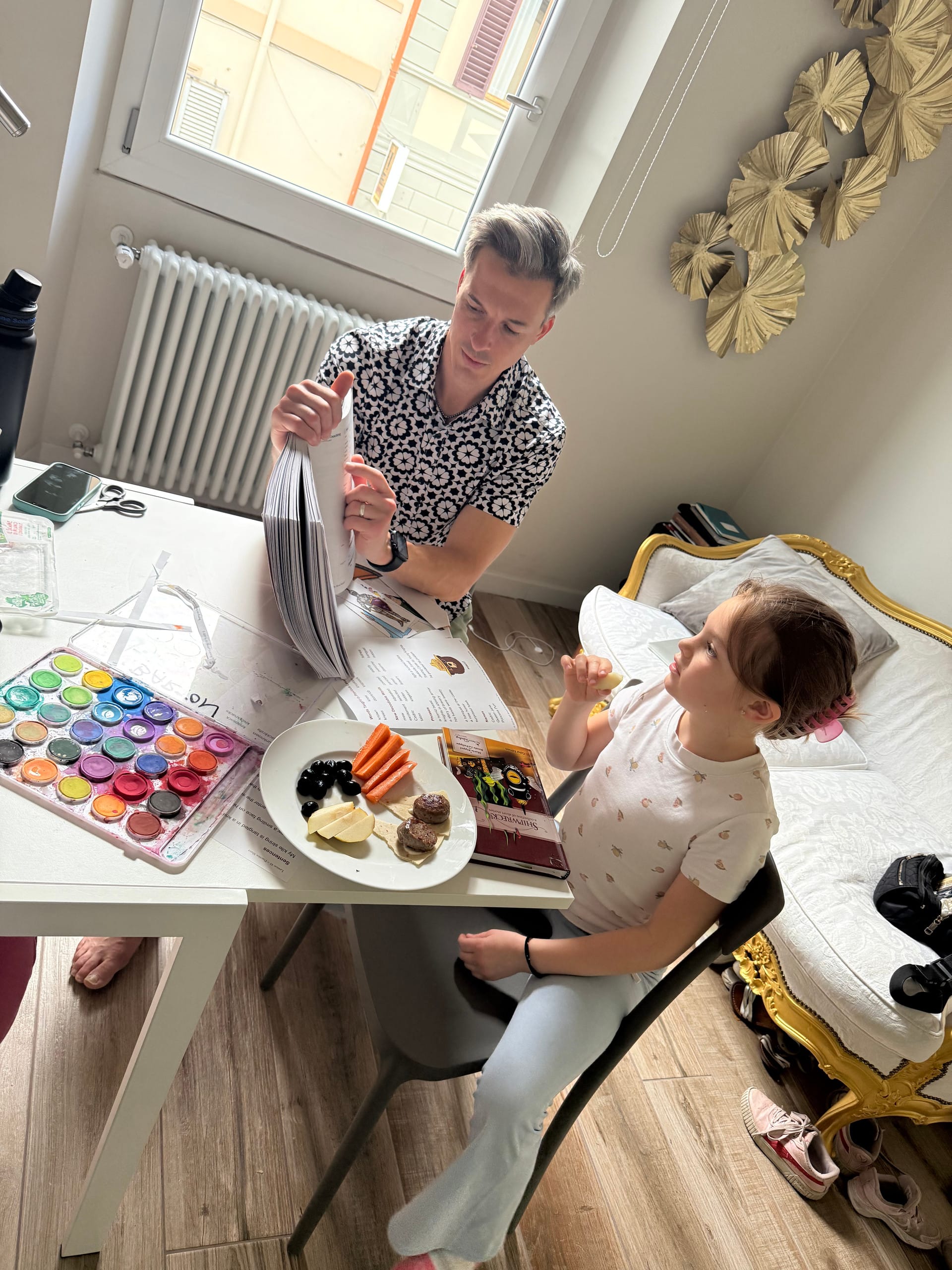 An adult flipping through a stack of practice worksheets at a kitchen table, a child seated alongside eating a snack and looking up, with a watercolor set and an open workbook on the table.