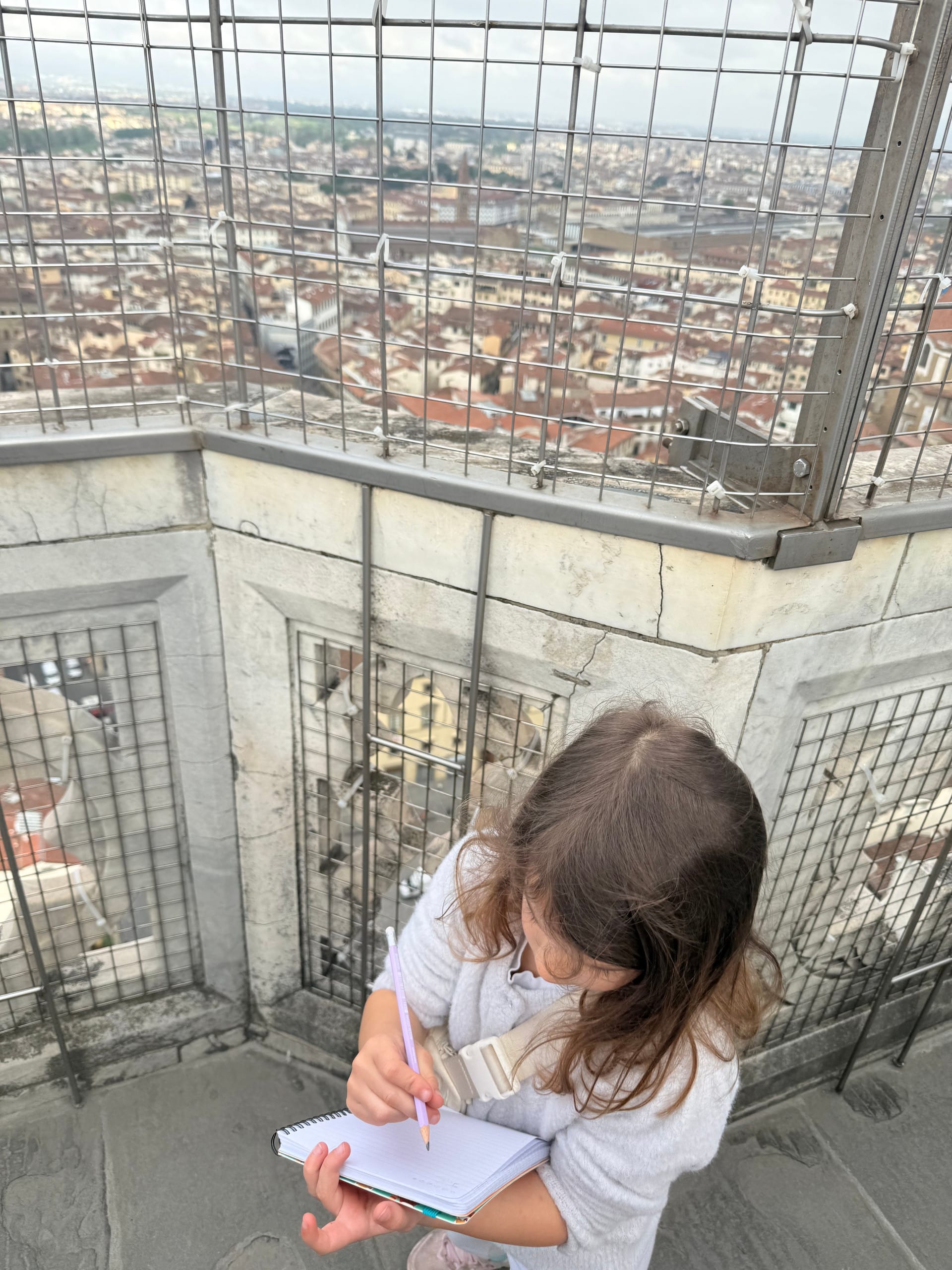 A child writing in a spiral notebook at the top of Giotto's Campanile, Florence rooftops visible through the safety mesh behind her.