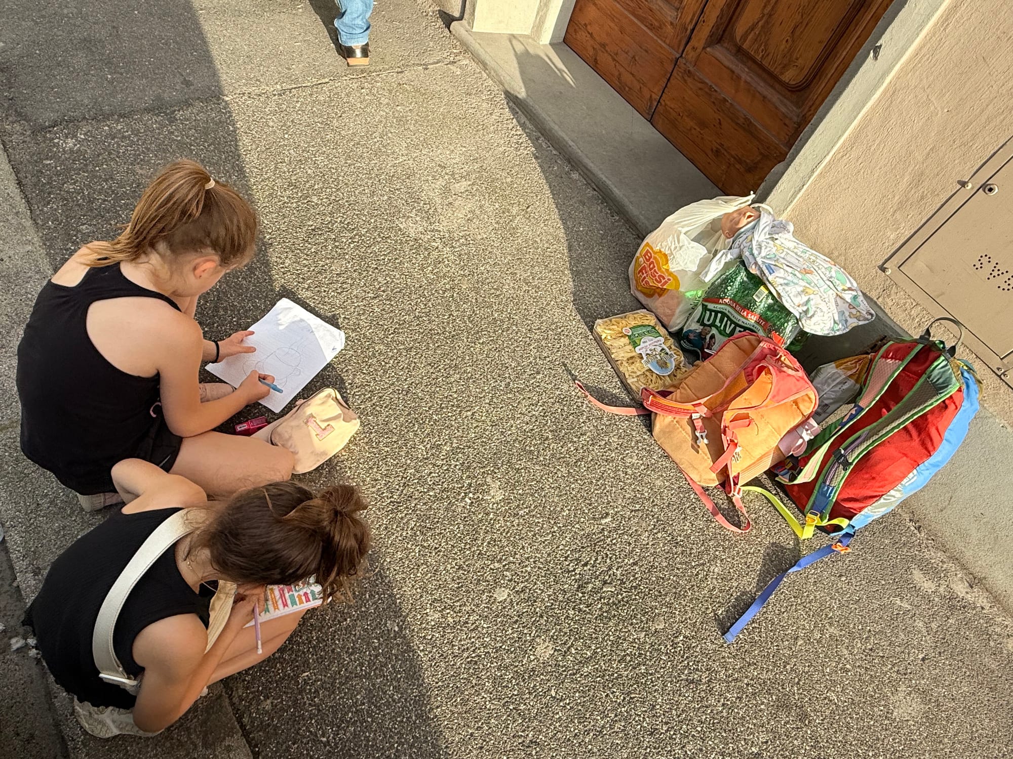 Two girls writing on paper on the pavement just outside their apartment door in Florence, shopping bags and backpacks set down beside them.