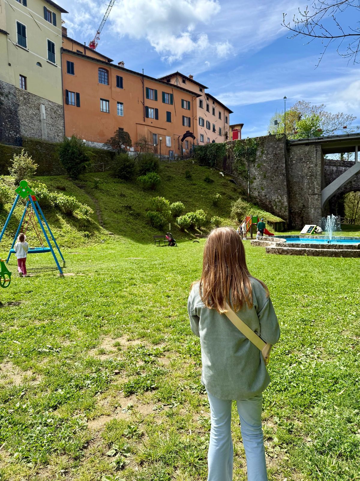 Lucy walking toward a running fountain in a valley playground in Barga, Italy, with orange buildings and a construction crane behind her