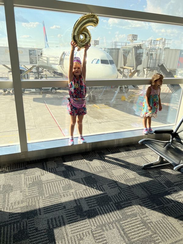 image alt	Lucy holding a gold number-6 balloon overhead at the airport gate, plane on the tarmac behind her