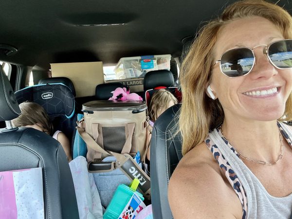 A packed SUV interior — bags, a cooler, and bins stacked to the ceiling — with two kids buckled in the back seat and a parent glancing back from the front.