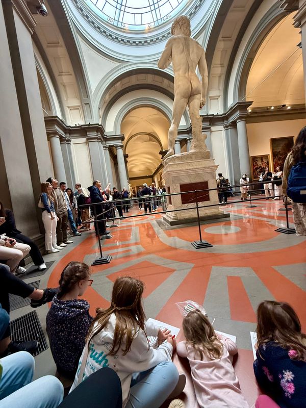Children coloring museum worksheets on the gallery floor, Michelangelo's David and other visitors in the background — the Accademia Gallery, Florence.