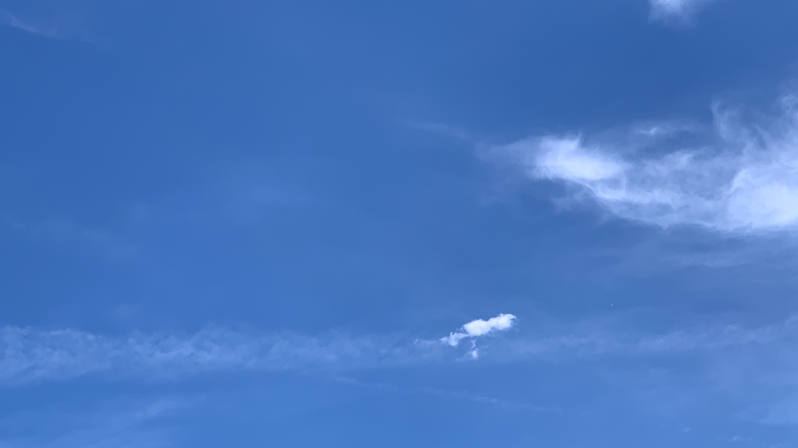 A patch of blue sky with some wispy cloud coming into the frame from the right, a fading contrail running across the lower part of the frame, and a tiny oblong brilliant white cloud perched on top of the contrail.