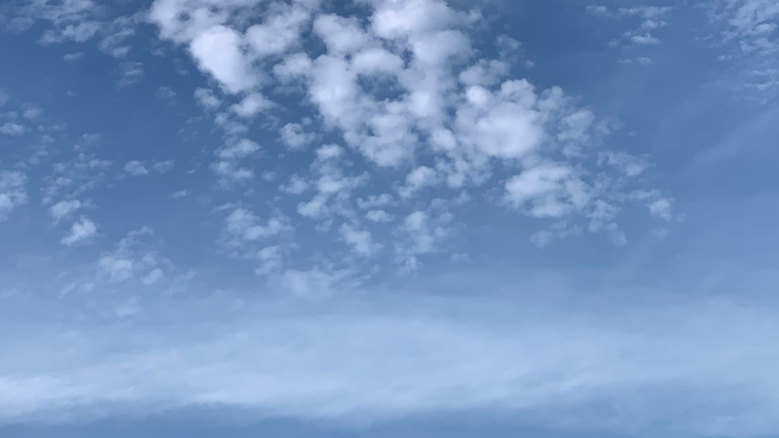 A patch of blue sky with a diffuse band of white running across the lower part of the frame and a cluster of white peppercorn clouds in the upper middle of the frame, their formation tipped toward the upper left.