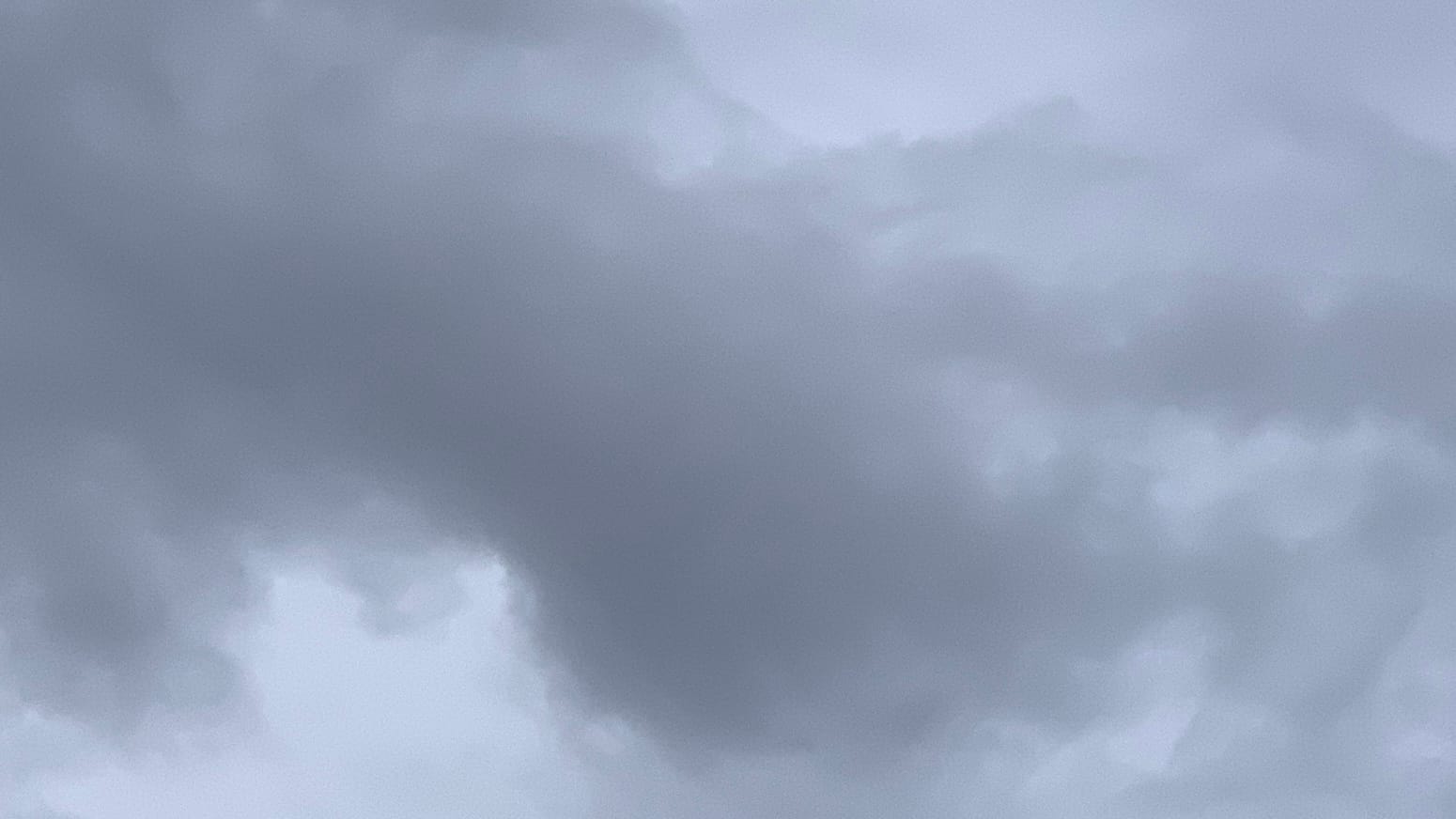 A patch of gray, cloudy sky with darker gray cloud shapes filling the middle of the frame and undulating across it. Faint tones of green and lilac are in the gray. 