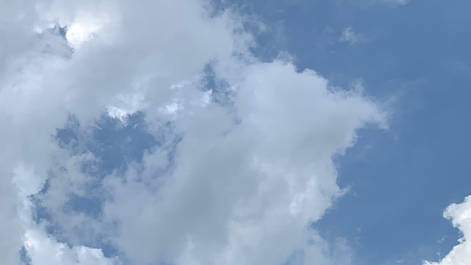 A patch of sky with clouds in tumult against a blue background. To the left and in the middle are pale gray clouds, coming apart to reveal bright white clouds and blue sky through the rifts. To the right is blue sky with pointy protuberances of white cloud poking in from the bottom right edge of the frame. 
