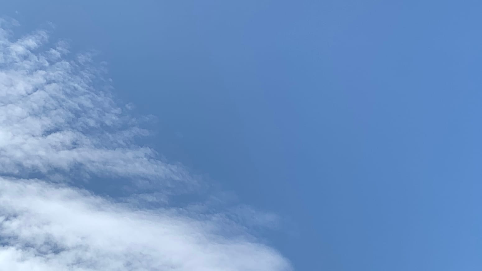 A patch of blue sky with the left side of the frame, angling down to the middle of the bottom, occupied by a sheet of thin white cloud separating into ridges. 