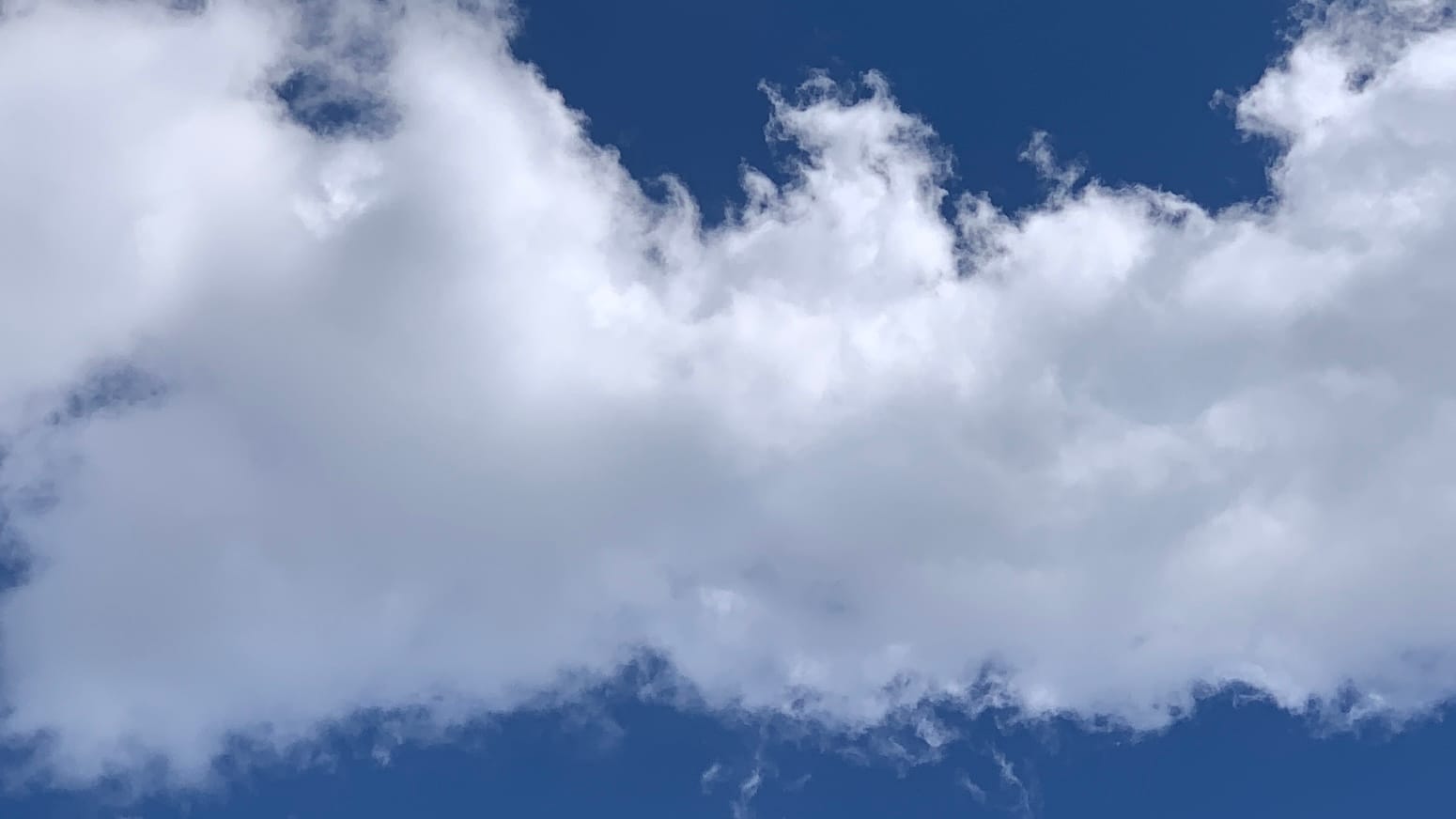 A patch of sky with a thick band of ragged-edged white cloud running through the middle of the frame, with a peak at the center, with deep blue sky at top and bottom