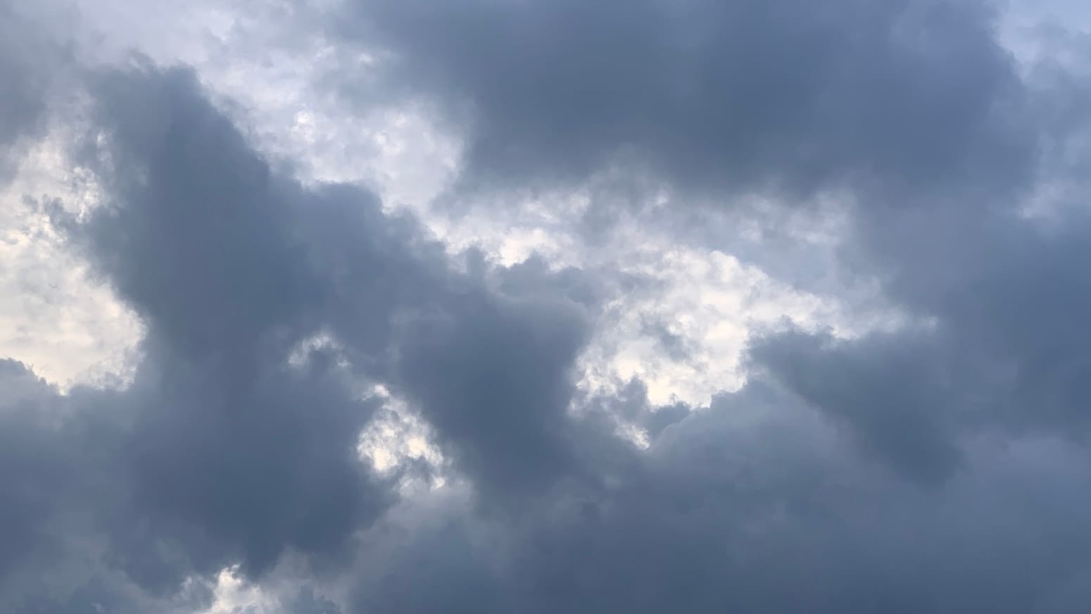 Layers of jagged dark-gray cumulus clouds with a bright hook-shaped negative space of lighter clouds reaching in among them from the upper left corner of the frame. 