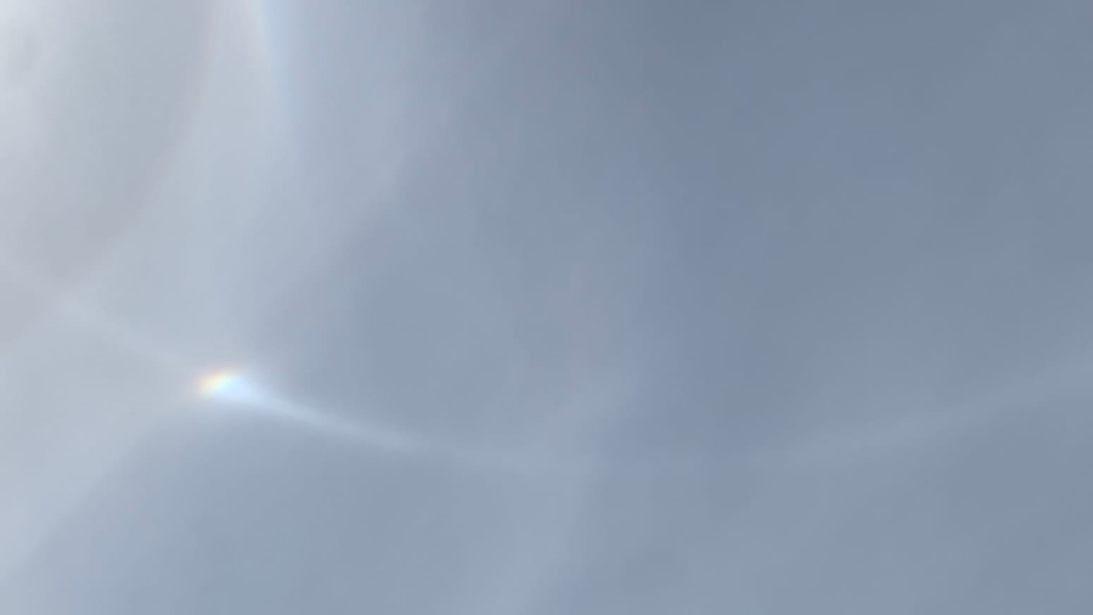A patch of bluish-gray sky with an arc of light, convex side down, bending across the frame, flaring toward the  left-hand side as it meets a sort of twisting pillar of refracted light. 