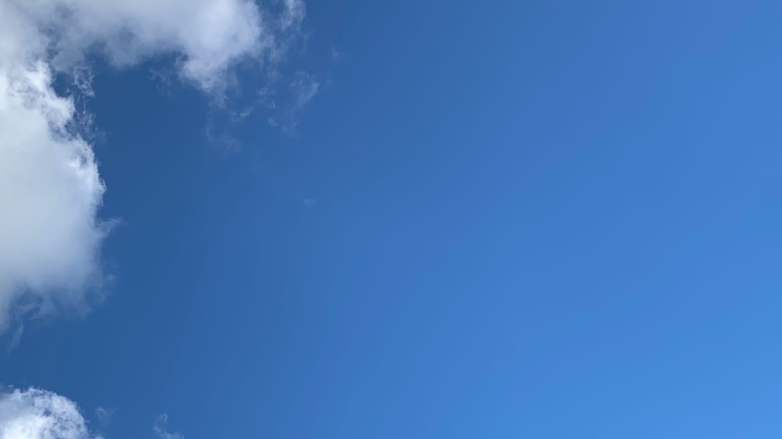 A patch of mostly blue sky with the edges of white cumulus clouds curling in from the top left and bulging in from the upper left and from the very bottom right corner, attended by a few loose wisps alongside.