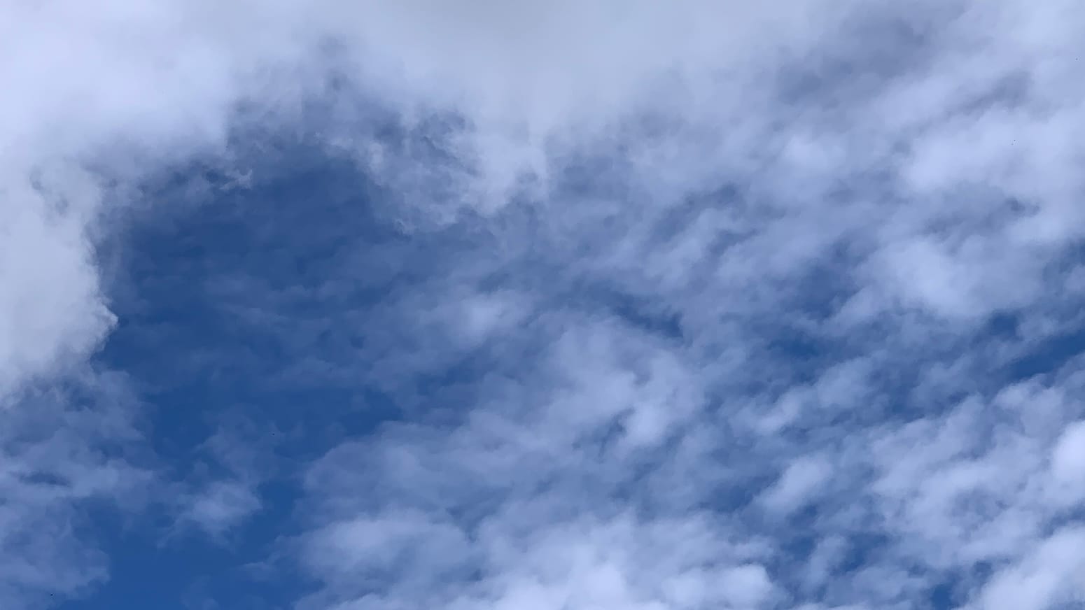 A patch of sky with loose white clouds letting blue show through, including a gap of almost open blue toward the upper left, framed there in the corner by some thicker white clouds. 