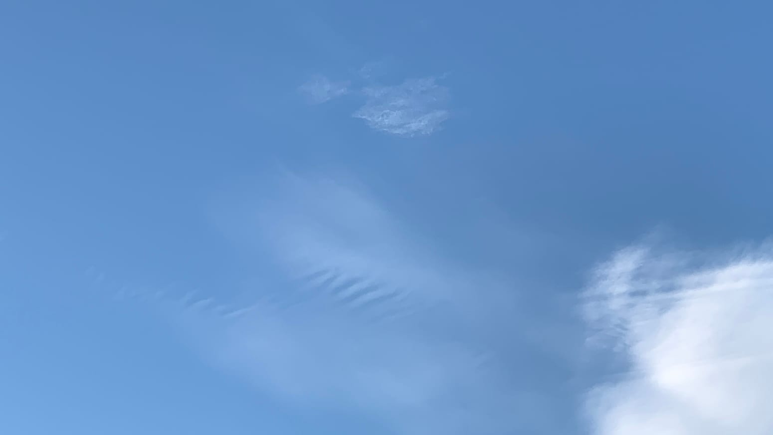 A patch of blue sky with some blurry and very transparent clouds in the center of the frame, and with some less transparent white cloud in the lower right corner. There are evenly spaced blue lines or furrows cutting through the middle of the transparent cloud, and there are straight lines of white and blue cutting through the top of the cloud in the corner. 