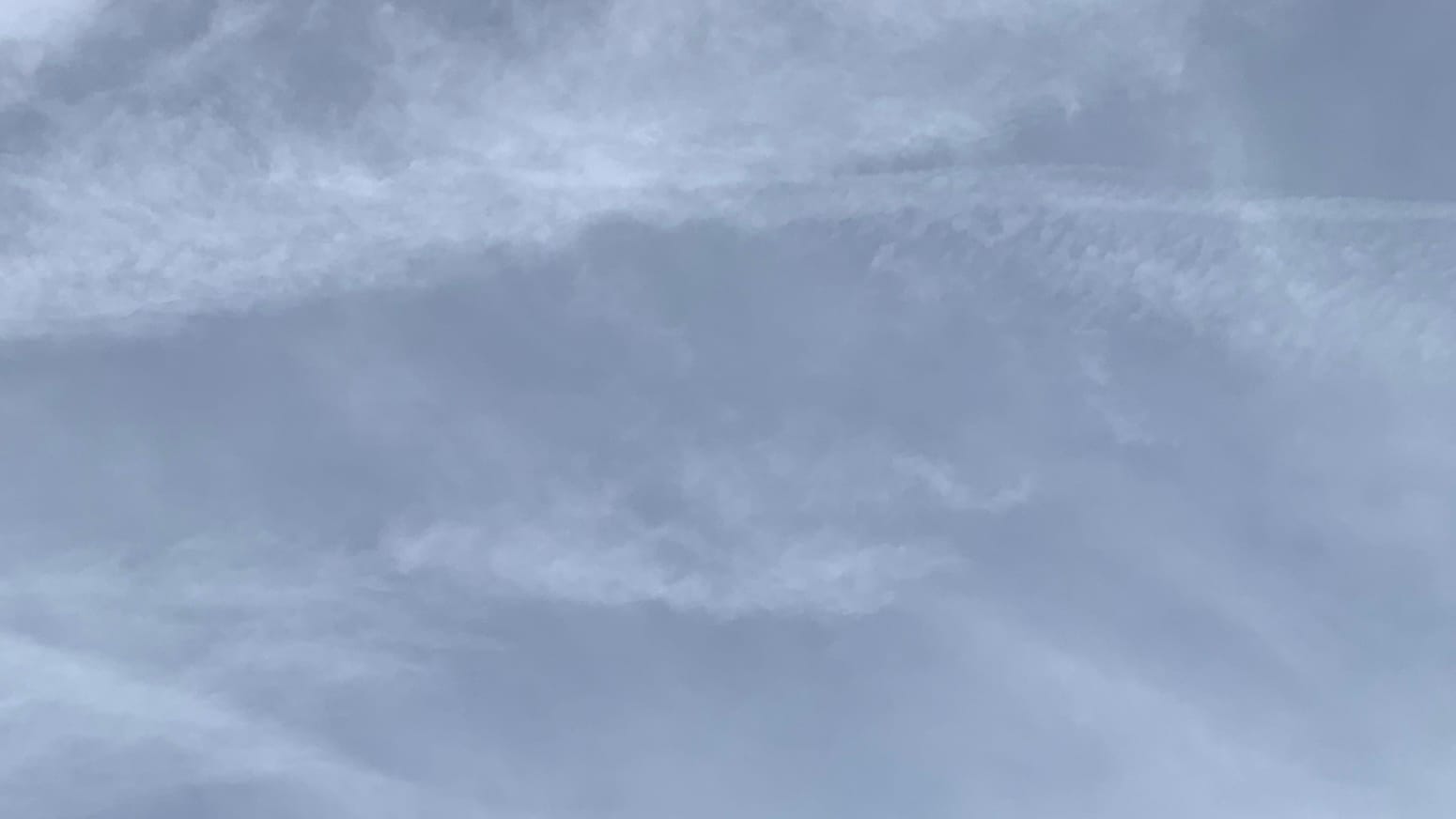 A patch of gray cloudy sky shot through with loose, intricately ruffled bits of more whitish gray clouds, including a fairly horizontal contrail, with even more transparent whitish gray clouds running on the diagonal behind. 
