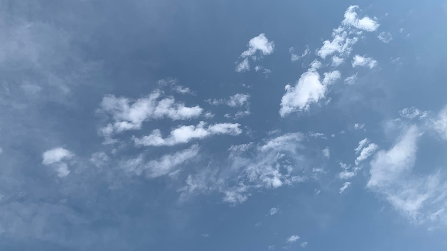 A patch of sky the faded color of old denim, with thin loose high clouds behind a spattering of small bright white clouds.