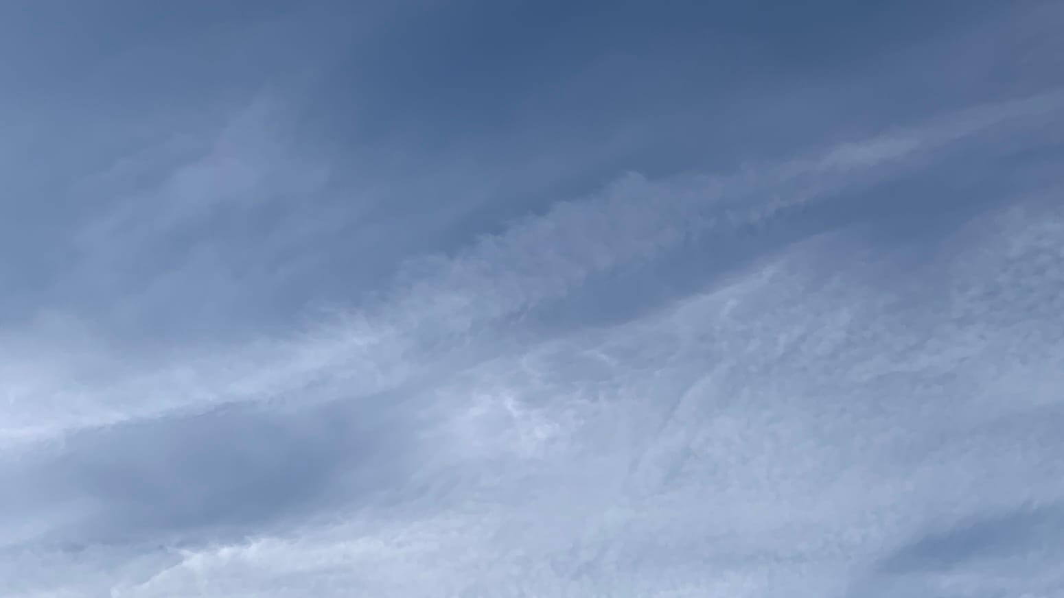 A sky of deep but desaturated blue, overlaid with the translucent remains of contrails, with an underlying grain angling up from the left side to the right. The contrail-generated cloud cover is thicker toward the bottom right and almost absent in the upper right and along the top of the frame.