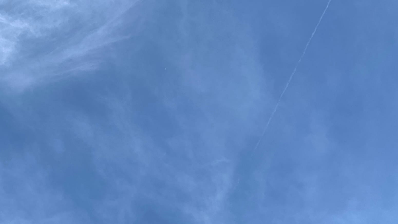 A patch of blue sky overlaid with a blurry network of faint, translucent clouds. The clouds are a bit denser in the top left corner. In the right half of the frame, where the sky is bluer, a thin and faint but very straight contrail runs about 30 degrees clockwise off the vertical.  
