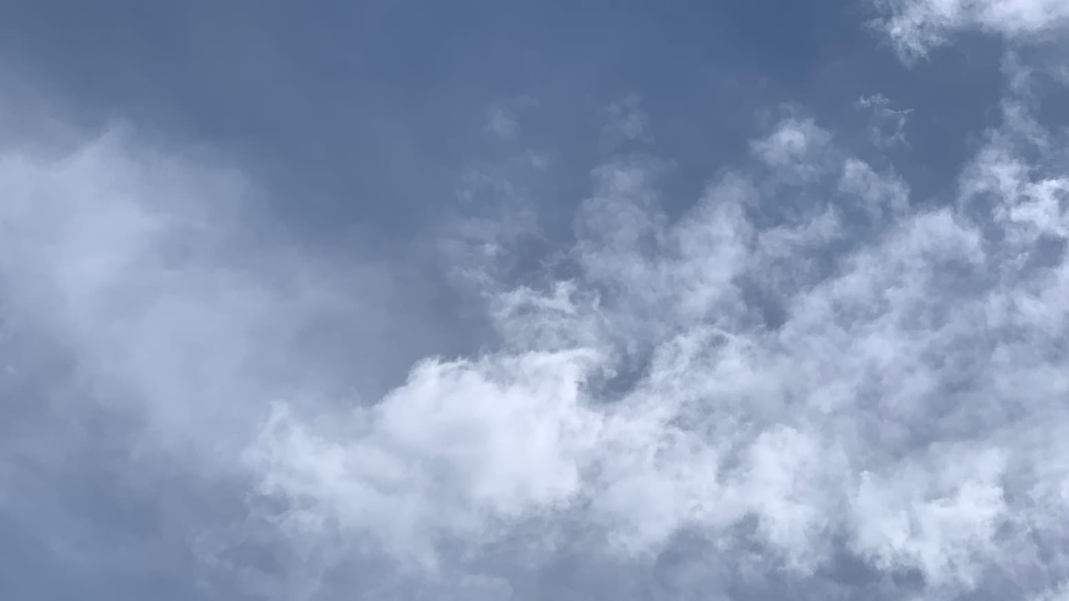 A patch of slatey blue sky with some blurry smears of thin cloud to the left and brighter, more distinct but loose-knit clouds, in clumps and wisps, taking up the lower right part of the frame and rising to curl like the crest of a wave beginning to break in the top right corner.