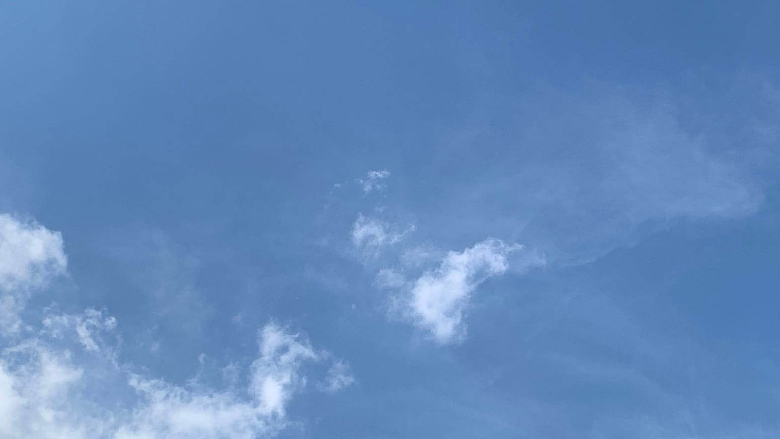 A patch of blue sky with white shreds of cloud in the lower left corner, a loose clump of shreds just below the center of the frame, and barely visible traces of spread-out cloud behind those. 