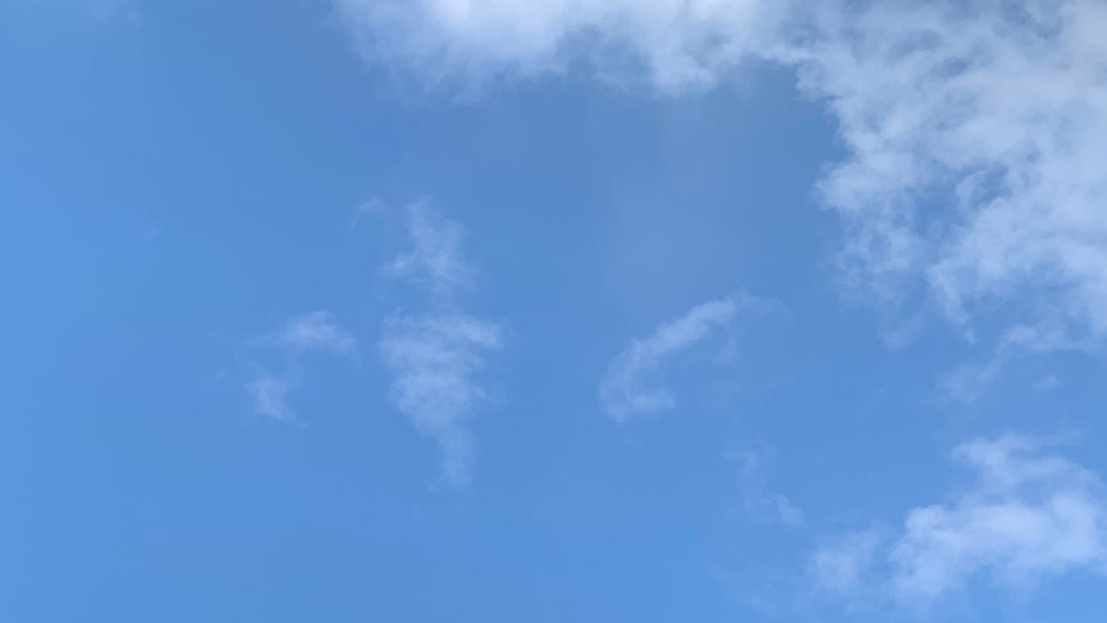 A path of light blue sky with thin shreds of cloud in the middle of the frame and somewhat denser but still thin cloud-matter wrapping up the right side of the frame and around the top right corner. 