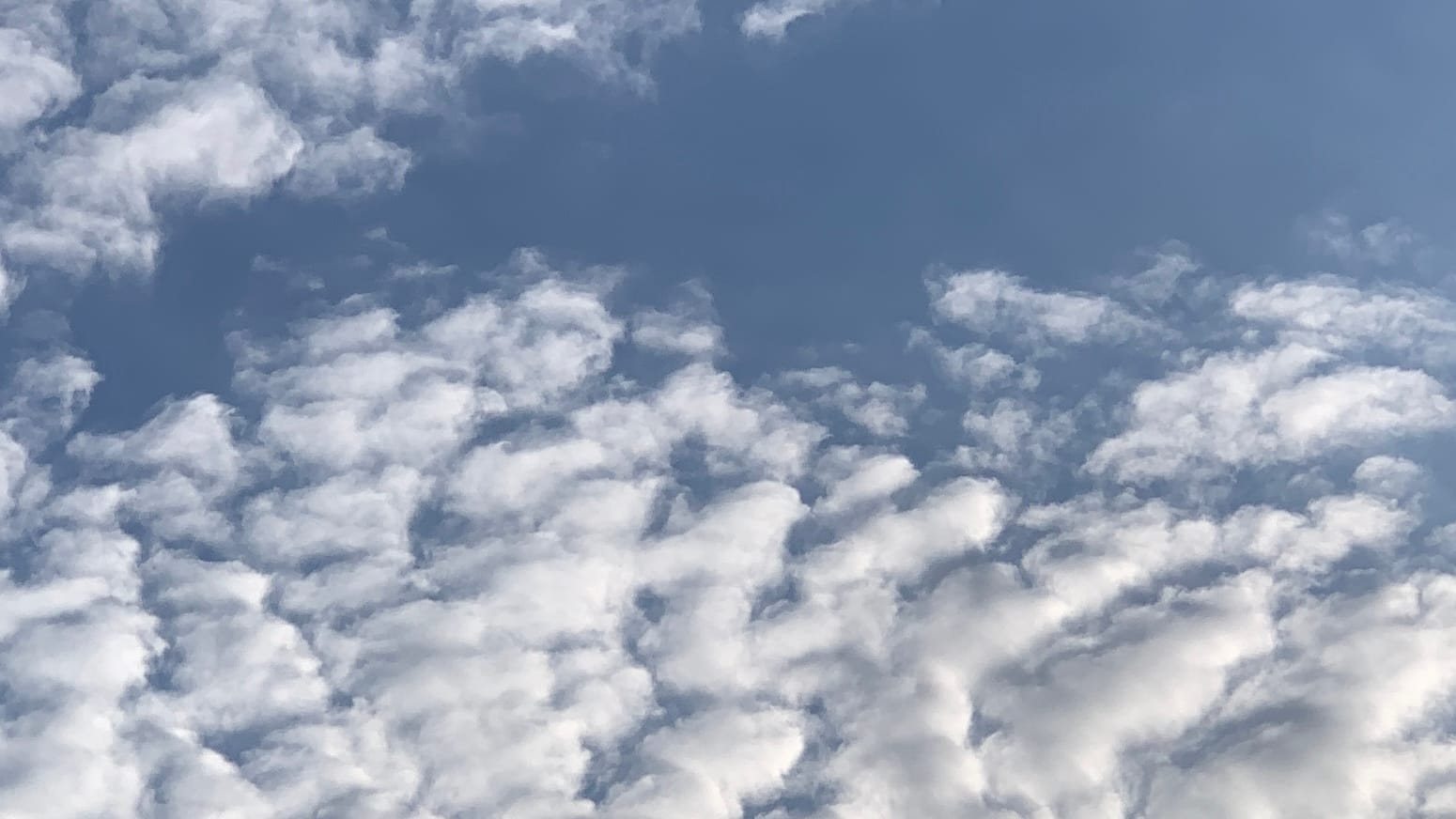 Shiny clustered fingerlike white clouds, with dark blue between them, stretching up toward a sort of open arm of blue reaching across the upper part of frame from the top right toward the left edge. In among all the other cloud forms a single wisp of white touching the center of the top of the frame is mirrored by a similarly shaped little rift of blue directly below it on the bottom of the frame.  