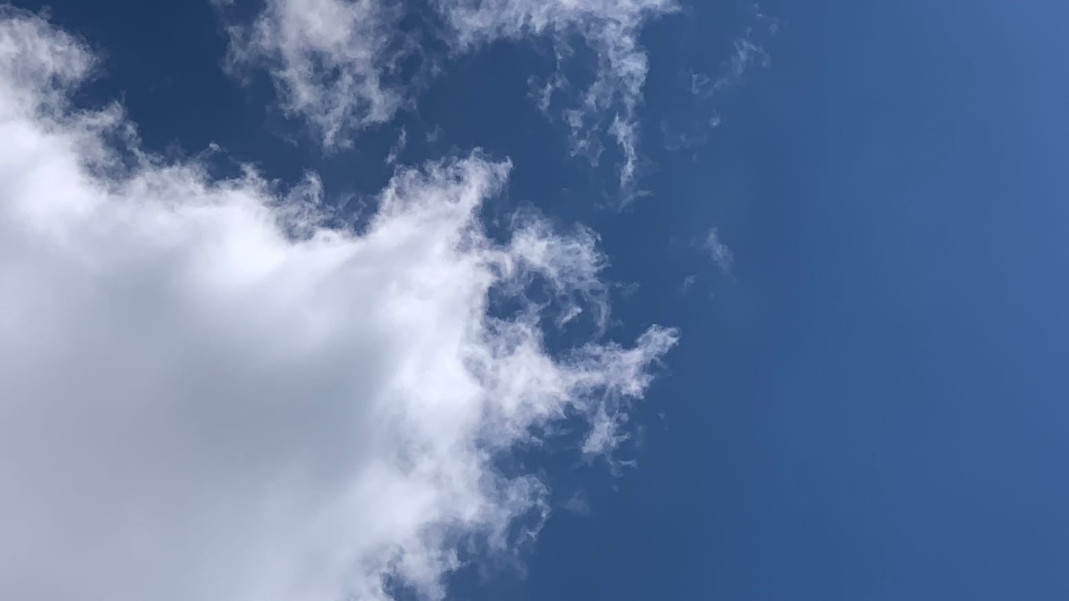 A patch of deep blue sky with a fluffy yet pointy white cloud jutting in from the bottom left corner to nearly fill the left edge of the frame. A shred of bonus cloud floats off the edge of it, tracing the shape of a promontory of the main cloud. 