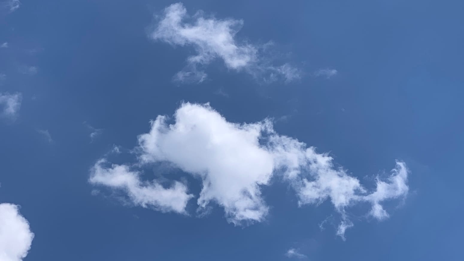 A white cloud with a hook-shaped appendage trailing off its right side floats in a fairly deep blue sky with a few other bits of cloud for company. 