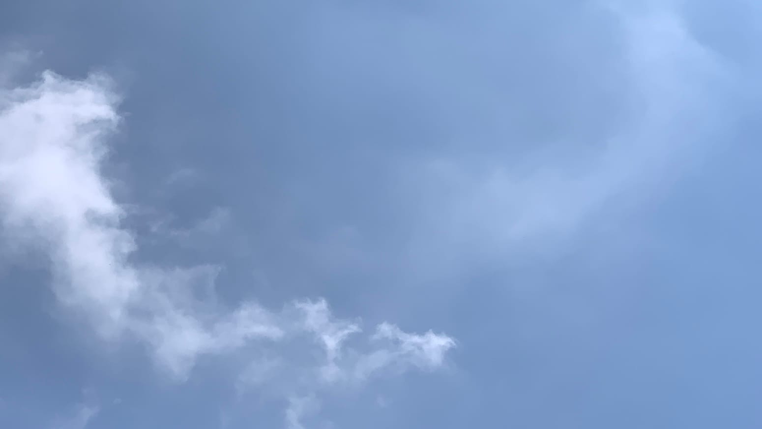 A patch of blue sky with a sort of jagged curve of white clouds curving from mid-upper left toward the bottom center of the frame, and a faint, smoother, translucent sort of mirror image of the curve, a little higher in the frame, curving from top right toward the center. 