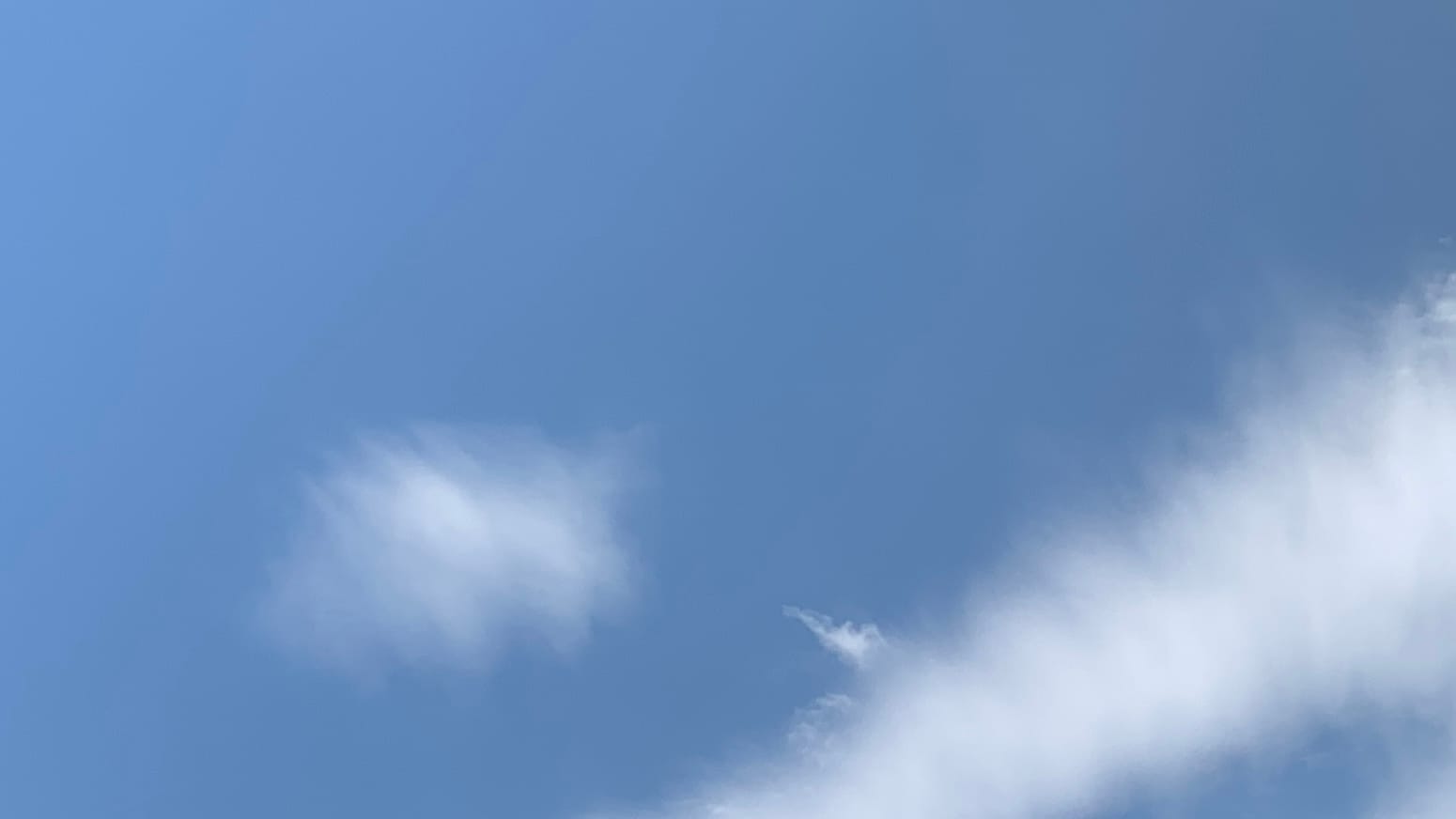 A patch of blue sky with smoothly blurry white clouds in it, apparently composed of little transverse cloud-parts jammed together. One is small and vaguely football-shaped and anther, crossing the lower right corner of the frame, looks like a piece of an elongated cone. Sticking out of that one, or emerging from behind it, is a much more sharply formed and denser-looking tiny horn of cloud. 