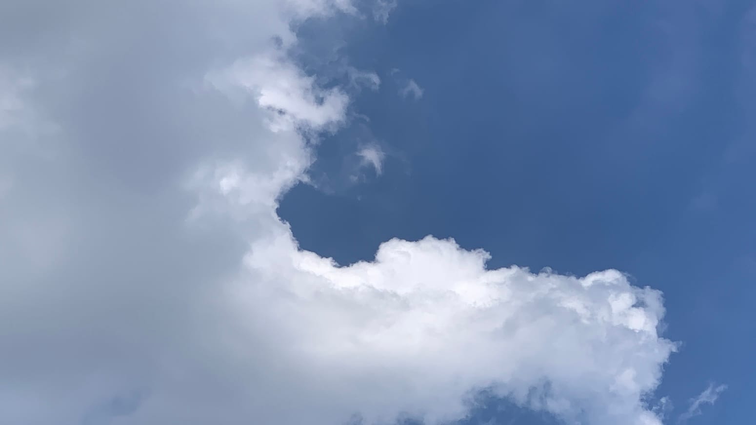 A patch of sky with deep blue in the upper right of the frame, with a few near-invisible blurry streaks of cloud on it, and a mass of pale gray and brilliant white cumulus filling the left side of the frame and extending a nearly right-angled arm most of the way across the bottom. 