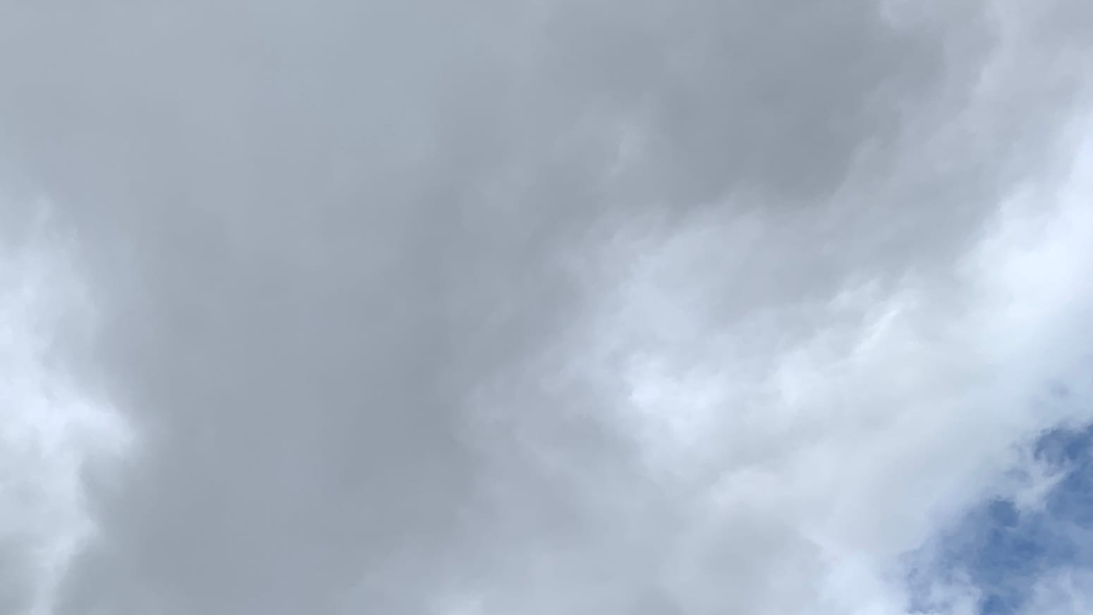 A patch of sky almost entirely filled with churning-looking clouds with a gray sort of hourglass pattern edged in white. The bottom right corner has blue showing through a webbing of thin white cloud. 