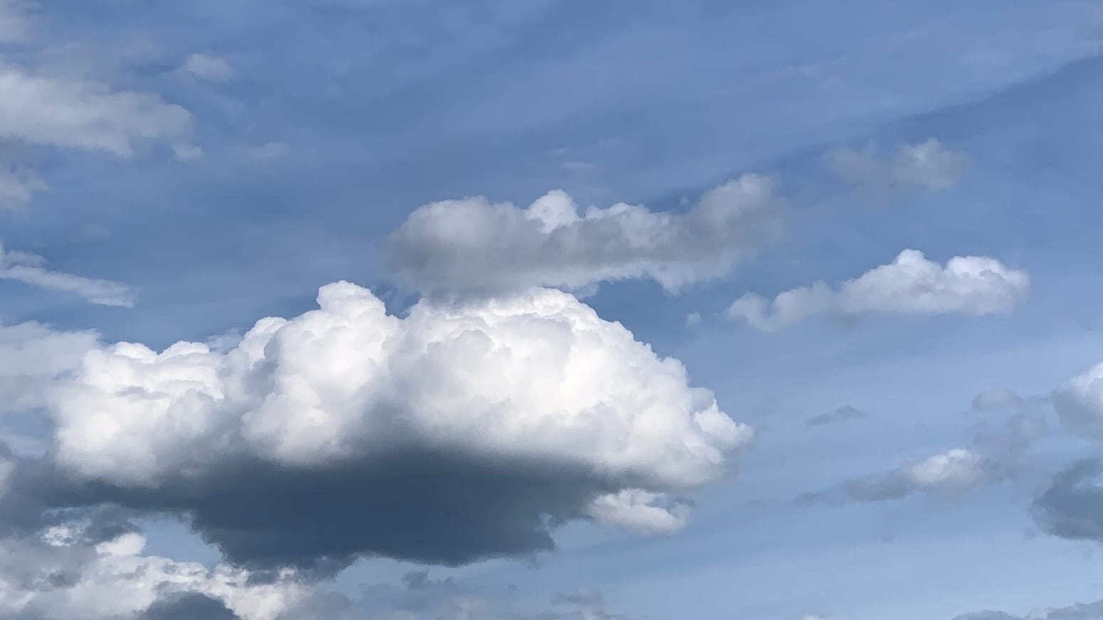 A white cumulus cloud, almost comically solid-looking with a flat shadow-blackened bottom, floats surrounded by less imposingly constructed squiggles of cumulus, some gray and some white and all very emphatically placed in three-dimensional space far below a translucent sheet of high white cloud that lets blue show through. The high cloud sheet has a straight-edge division, running on a flattened diagonal, separating the whiter and bluer parts of the sky. 