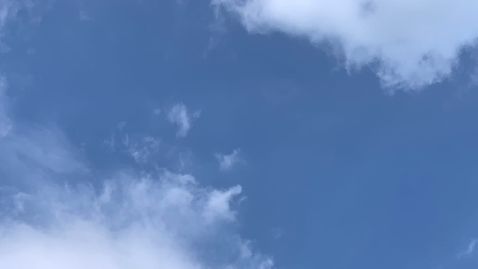 A patch of blue sky with loose white clouds reaching into the frame from the bottom left and top right, like the banks of a broad and meandering blue river. 